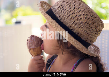 Petite fille avec un chapeau de paille et une robe d'été bénéficie de la chaleur de l'été manger un cône rafraîchissante de crème glacée au chocolat, il fond dans sa main alors qu'elle Banque D'Images