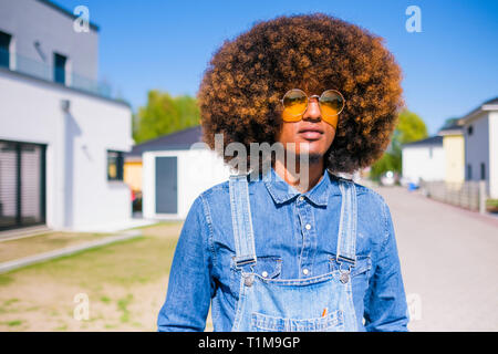 Portrait cool jeune homme avec afro dans des combinaisons en denim debout sur la route ensoleillée Banque D'Images