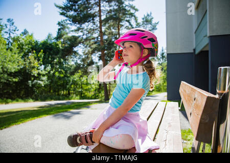 Petite fille dans helmet assis sur un banc Playing with toy téléphone cellulaire. Journée ensoleillée en banlieue. Enfance heureuse. Banque D'Images