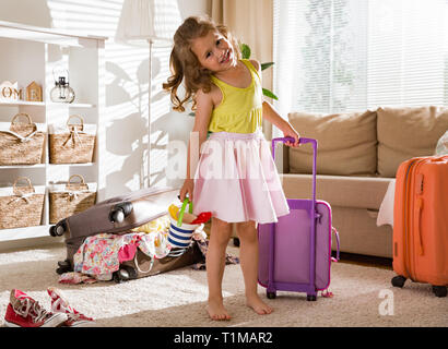 Heureuse petite fille assise dans la salle de séjour à la maison, soigneusement l'emballage des vêtements d'été colorés et des jouets en assurance valise pour un nouveau voyage. T de l'enfant Banque D'Images