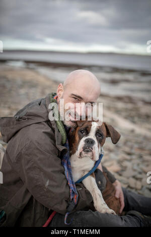 Une vue avant-shot of a mature man assis sur une plage avec son chien boxer blanc et brun, ils adoptent l'autre sur la journée dans le froid Banque D'Images