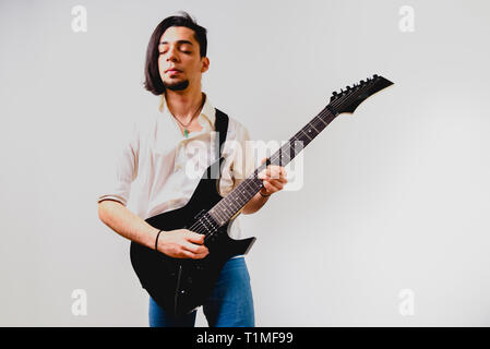 Jeune guitariste aux cheveux longs noir excité jouant de sa guitare électrique, isolé sur fond blanc. Banque D'Images