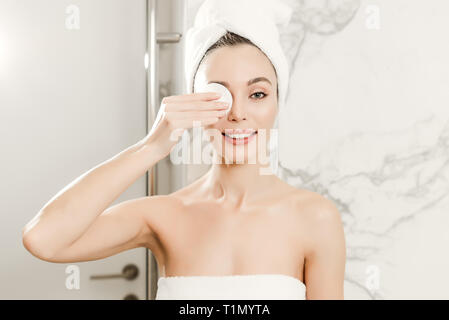 Belle jeune femme enveloppée dans des serviettes nettoie son visage avec un coton dans la salle de bains. maquillage et soins de beauté concept Banque D'Images