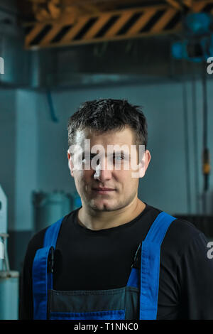 Portrait d'un jeune homme séduisant d'apparence de race blanche dans un atelier de métallurgie. L'homme en salopette de travail se penche sur l'appareil photo. Banque D'Images