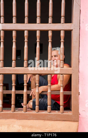 Portrait d'un homme cubain, à Trinidad, Sancti Spiritus, Cuba. Scène de rue montrant l'homme se détendre derrière un écran de sécurité en bois peint Banque D'Images