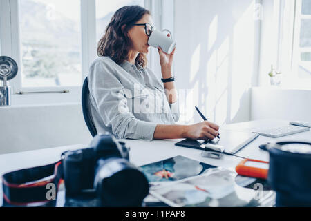 Femme artiste de boire du café tout en travaillant au bureau. Femme photographe photos retouche dans Office et de boire une tasse de café. Banque D'Images