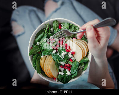 Femme en jeans holding vegan saladier avec les épinards, poire, pomme grenade, fromage. Le petit-déjeuner végétarien, végétalien, régime alimentaire concept. Jeune fille en jeans avec fourche holding genoux et mains visible Banque D'Images