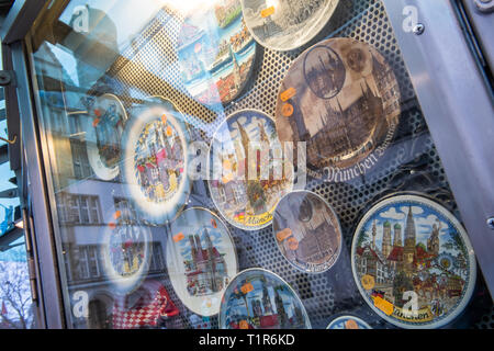 27 mars 2019, Bavaria, Munich : plaques de décoration avec des motifs de la capitale bavaroise accrocher dans un magasin de souvenirs dans la zone piétonne du centre-ville. La vitrine reflète les tours de la Frauenkirche. Photo : Peter Kneffel/dpa Banque D'Images