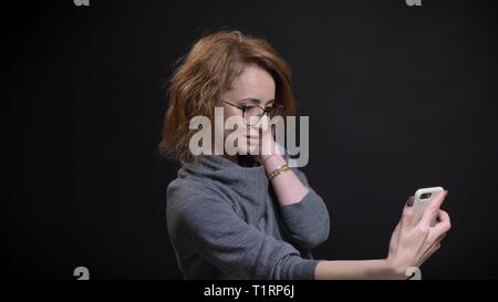 Closeup portrait of middle-aged femme aux cheveux rouge extravagant dans les verres en tenant vos autoportraits sur le téléphone et posant devant l'appareil photo Banque D'Images