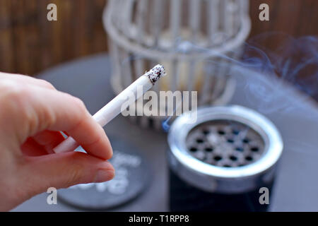 Close up of a woman's hand holding a cigarette allumée avec cendrier floue sur la table en arrière-plan Banque D'Images