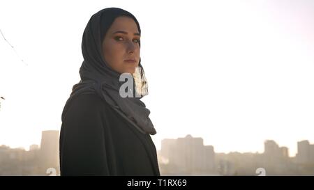 De jeunes pousses Gros plan jolie silhouette féminine en hijab à tout droit à l'appareil photo s'est tourné vers le côté avec urban city sur l'arrière-plan Banque D'Images