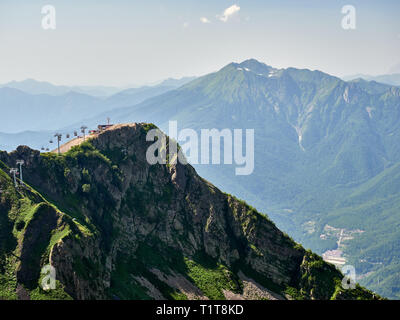 Les pics d'une haute montagne pente avec cable car, en face de laquelle est une vallée avec des maisons d'habitation. Ciel bleu dans les montagnes un jour d'été. K Banque D'Images