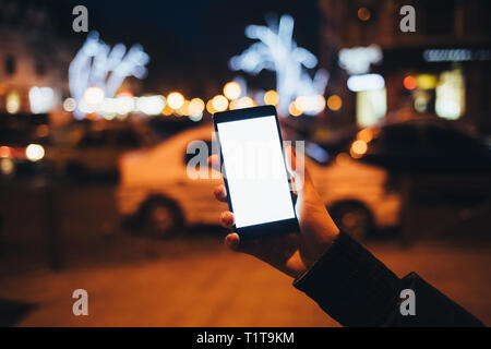 Femme's hand holding montrant smart phone avec écran vide près de road et de voitures sur rue en ville la nuit, les lumières brillantes à l'arrière-plan. Banque D'Images