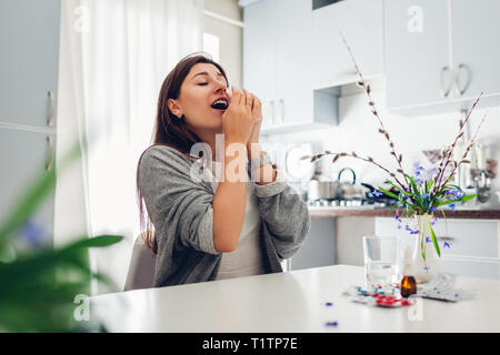 Allergies de printemps. Jeune femme éternuements à cause de fleurs entouré de comprimés sur la cuisine à la maison. Concept d'allergies saisonnières. Banque D'Images