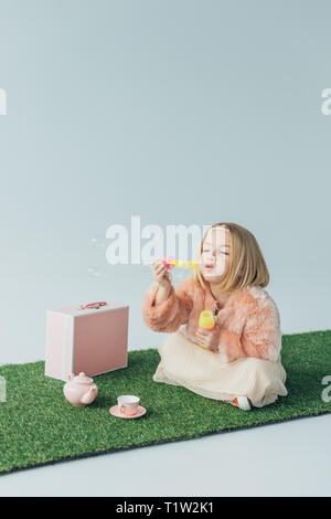 Cute kid avec jambes croisées assis sur un tapis d'herbe et en soufflant des bulles de savon isolé sur gray Banque D'Images