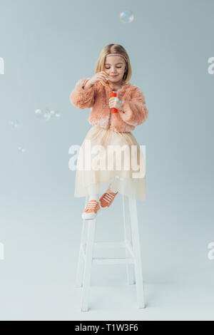 Adorable gamine dans un manteau en fausse fourrure et jupe assise sur une chaise haute et soufflant des bulles de savon Banque D'Images