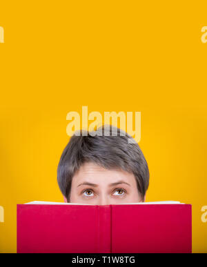 Jeune femme book reader se cache derrière un livre rouge, à la recherche jusqu'a l'expression réfléchie face au mur jaune isolé avec copie espace. La fema curieux Banque D'Images