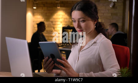 De jeunes pousses Gros plan pretty caucasian businesswoman en utilisant la tablette alors qu'il était assis en face de l'ordinateur portable à l'intérieur sur le lieu de travail Banque D'Images