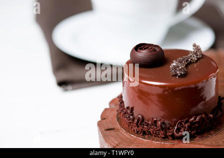 Un délicieux gâteau au chocolat avec des morceaux de chocolat repose sur un support en bois à côté d'une tasse blanche, qui se dresse sur une table en bois blanc Banque D'Images