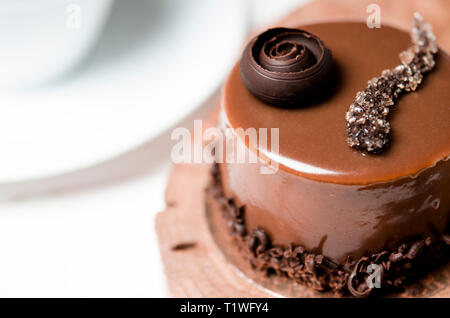 Un délicieux gâteau au chocolat avec des morceaux de chocolat repose sur un support en bois à côté d'une tasse blanche, qui se dresse sur une table en bois blanc Banque D'Images