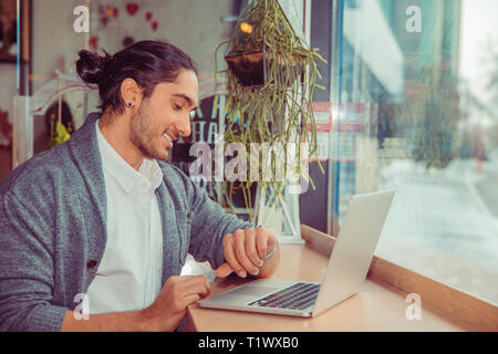 Homme heureux souriant à la montre-bracelet à tout en travaillant à l'ordinateur. Closeup portrait of a guy in casual wear assis près de fenêtre à table dans la vie Banque D'Images