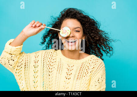 Happy african american girl smiling bouclés tout en couvrant avec des yeux bleu isolé sur lollipop Banque D'Images