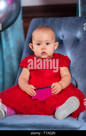 Petit enfant fille en robe rouge de fête avec le smartphone dans ses mains, assis sur une chaise bleue et à l'avant avec l'impassible visage mignon Banque D'Images