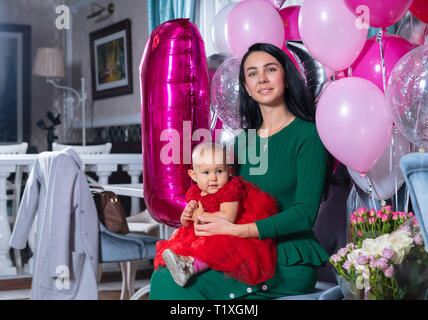 Un premier anniversaire fête de la mère avec sa fille posant avec la fille de bébé sur ses genoux entourés par des ballons et des fleurs rose partie Banque D'Images