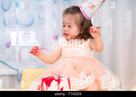 Petite fille profiter de son anniversaire assis devant le gâteau avec les mains et le visage portant un chapeau et robe rose Banque D'Images