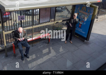 Un vieux monsieur essuie ses lunettes avec un chiffon propre, comme une dame pose la sienne pour voir les bus venant en sens inverse, sur le Danemark Hill à Southwark, le 28 mars 2019, à Londres, en Angleterre Banque D'Images