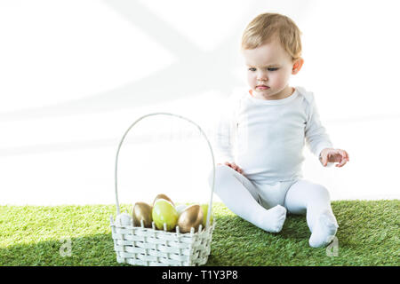 Adorable enfant assis sur l'herbe verte près de panier avec de la paille d'or et jaune des oeufs de Pâques isolated on white Banque D'Images