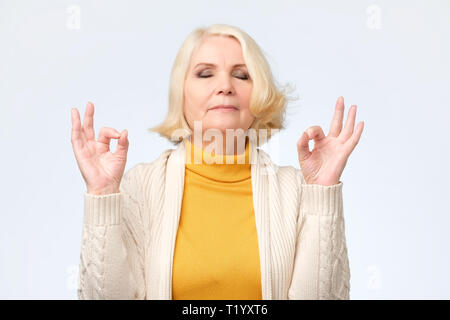 Senior woman smiling with eyes closed faisant le geste de la méditation avec les doigts. Concept de yoga. Banque D'Images