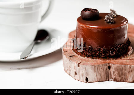 Un délicieux gâteau au chocolat avec des morceaux de chocolat repose sur un support en bois à côté d'une tasse blanche, qui se dresse sur une table en bois blanc Banque D'Images