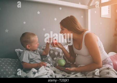 Mère enceinte et son petit garçon sont de manger une pomme et pêche dans le lit t accueil le matin. Style décontracté dans la chambre Banque D'Images