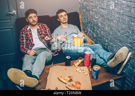 Deux mecs assis sur le canapé et la tenue de la nourriture et des boissons dans leurs mains. Ils se sentent détendus et fatiguée après un long match jouer différents jeux. Banque D'Images