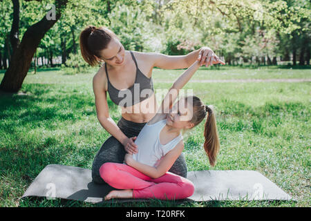 Jeune femme aide sa fille à s'étirer. Elle est assise derrière elle et tenant sa main dans l'air tout en poussant sur la hanche de la jeune fille. Petite fille est sitti Banque D'Images