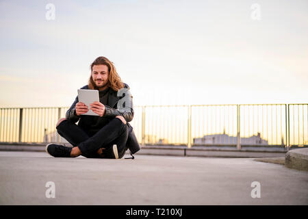 Portrait de jeune homme barbu assis sur niveau parking au crépuscule using digital tablet Banque D'Images