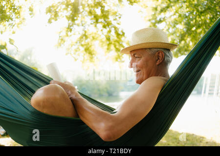 Senior man wearing straw hat relaxing in hammock au lakeshore reading book Banque D'Images