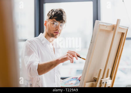 Focus sélectif de handsome artiste en verres looking at camera pendant le dessin sur toile Banque D'Images