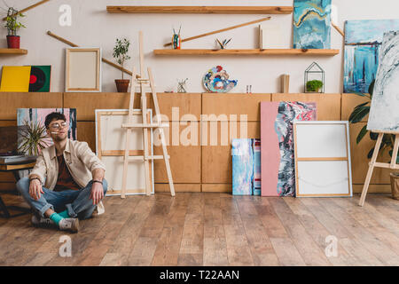 Artiste en jeans et chemise rose assis sur le sol dans un grand studio de peinture et cigarette Banque D'Images