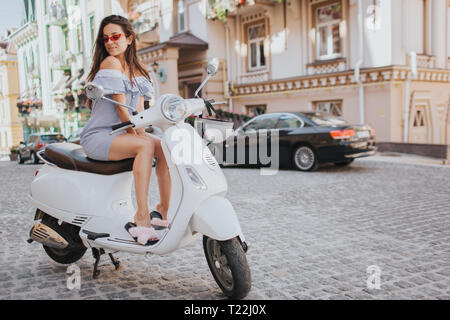 Fabulous girl est assis sur moto blanc et regardant vers le bas. Elle pose sur l'appareil-photo. Elle est debout au milieu de la route Banque D'Images