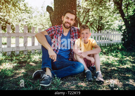 Belle photo de père et fils assis ensemble sous arbre et à la recherche sur l'appareil-photo. Ils sont souriants. Adulte est hugging enfant Banque D'Images