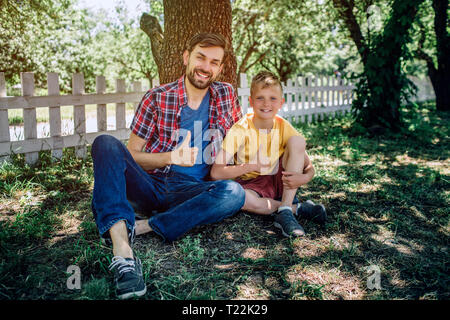 Bel homme et l'enfant sont assis côte à côte sur l'herbe et à la recherche sur l'appareil-photo. Ils sont souriants et à la recherche sur l'appareil-photo. Les garçons tiennent leur grand bravo Banque D'Images