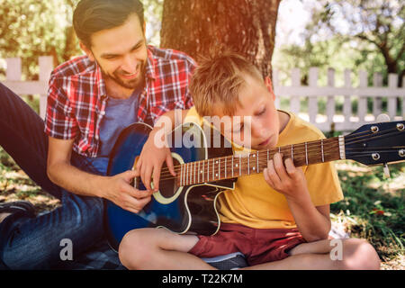 Garçon talentueux est à la recherche sur les cordes et à jouer de la guitare. Son père est assis en dehors de lui et l'aider. Adulte est smiling Banque D'Images