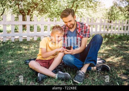 L'homme et l'enfant sont assis ensemble sur l'herbe avec leurs jambes croisées et à la recherche au téléphone avec intérêt et d'émerveillement. Ils sont en train de jouer avec elle. Regardez les gars Banque D'Images