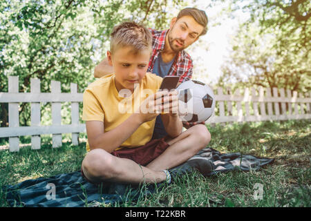 Homme barbu est assis à l'arrière de son fils, il veut jouer au jeu footbll avec enfant. Kid n'est pas intéressé par cela. Il joue avec le téléphone Banque D'Images