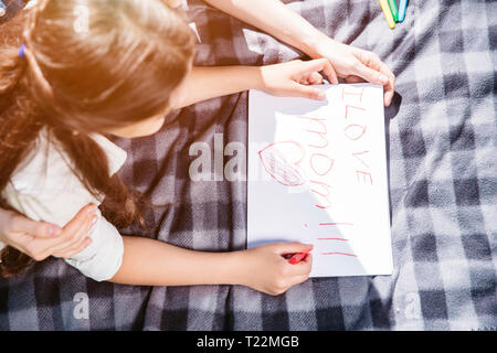 Sweet et belle photo de girl lying on blanket et de dessin. Elle est à l'aide de crayon de couleur rouge. Elle a écrit sur un morceau de papier pf J'aime maman Banque D'Images