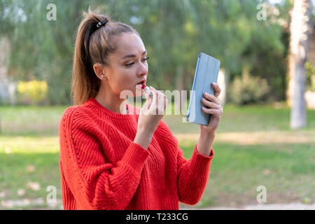 Young woman applying lipstick dans apark, using digital tablet comme un miroir Banque D'Images