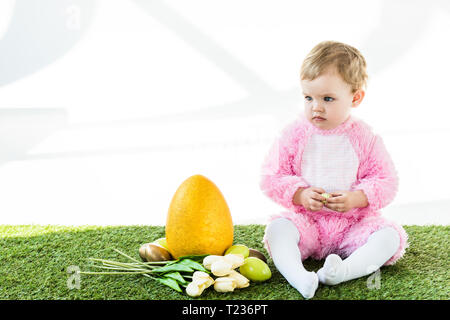 Adorable enfant en costume rose fluffy assis près de l'œuf d'autruche jaune coloré, les oeufs de poule et tulipes isolated on white Banque D'Images