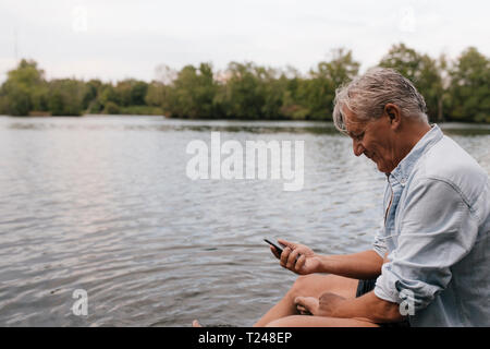 Senior man sitting at a lake using cell phone Banque D'Images
